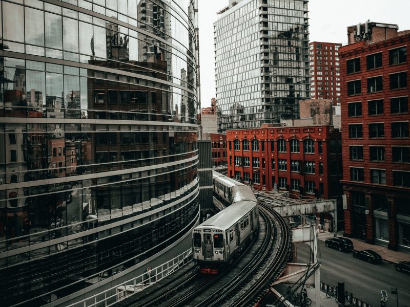 An aerial view of a train passing by on West Kinzie Street in Chicago. The city is reflected in a nearby building's windows.